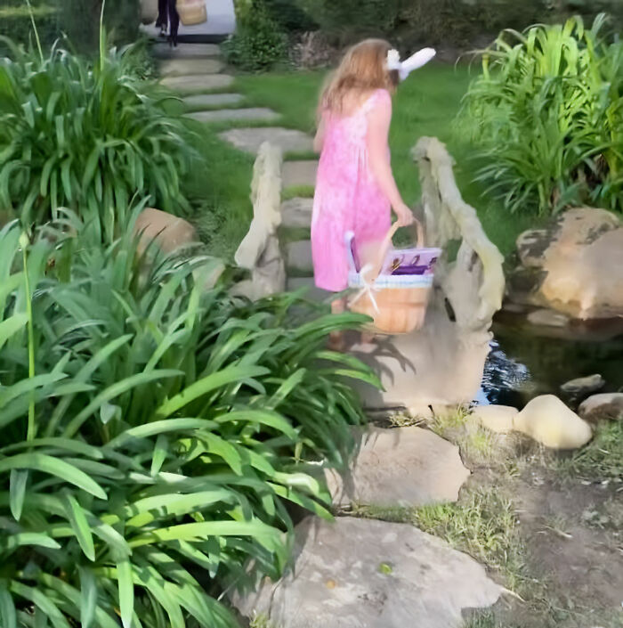 Child wearing bunny ears carrying Easter basket walking over stone bridge in garden, Meghan Markle Easter clip sparks backlash.