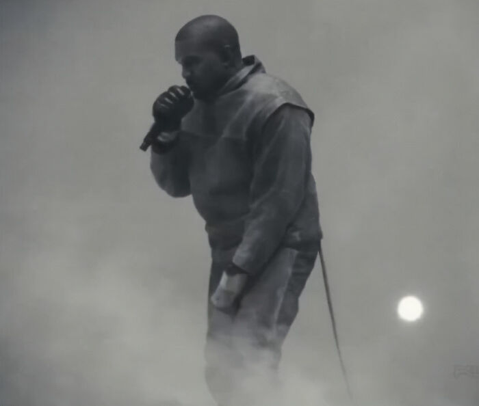 Kanye West se apresentando no palco com microfone em meio a uma atmosfera enfumaçada em um evento de música ao vivo.