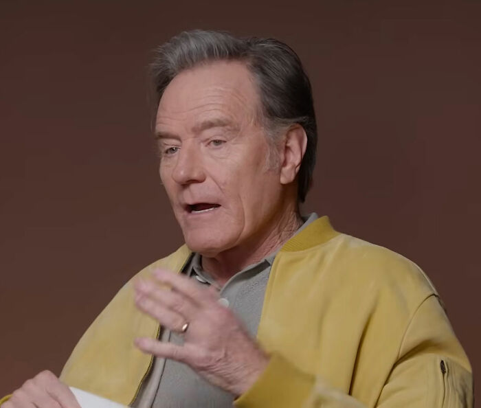 Bryan Cranston speaking with expressive hand gestures, wearing a yellow jacket in an indoor interview setting.