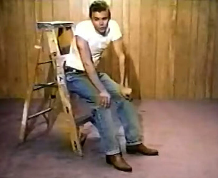 A man in a white T-shirt and jeans sitting on a wooden ladder in a plain room showing PR mistakes concept.