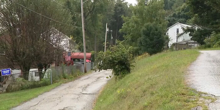 Uma estrada rural com colinas verdes e casas, uma delas com a bandeira de Trump, relacionada ao desaparecimento de uma criança de 9 anos.