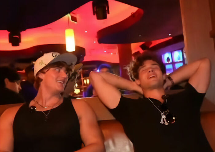 Two young men at a bar with red lighting, one wearing a white cap, the other relaxing with hands behind head &ndash; looksmaxxer livestream.