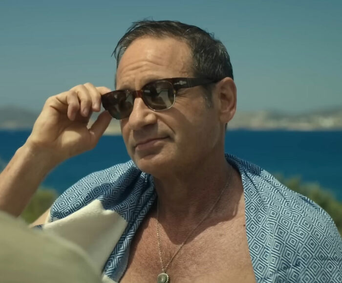 Homem ajustando óculos escuros à beira-mar, usando xale estampado, sem relação com estrela de TV dos anos 90, de saia e peruca loira atirando.