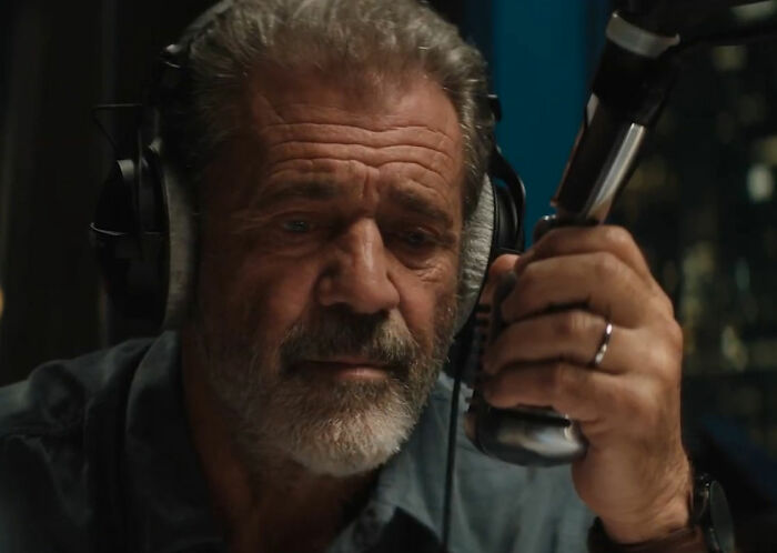 Mel Gibson com barba grisalha e fones de ouvido, segurando um microfone. Seu amor secreto.