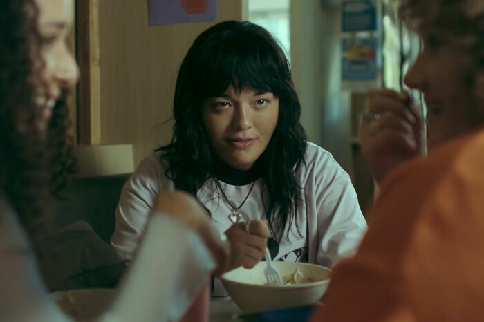 A young person with dark hair looks intently while eating from a bowl with a fork, amidst other people. Horror movie.