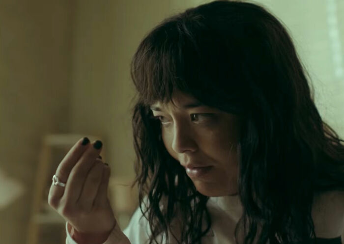 A woman with dark hair and black nail polish holds something tiny, staring intently, reflecting the horror movie's theme of being thinner.