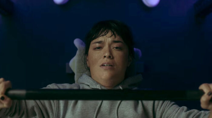 A woman in a grey hoodie strains while bench pressing, her face showing effort. The image hints at the horror movie condemning the thinner trend.