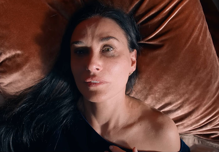 A woman with dark hair and wide, anxious eyes, lying on a brown velvet pillow, looking terrified in a horror movie about being thinner than ever.