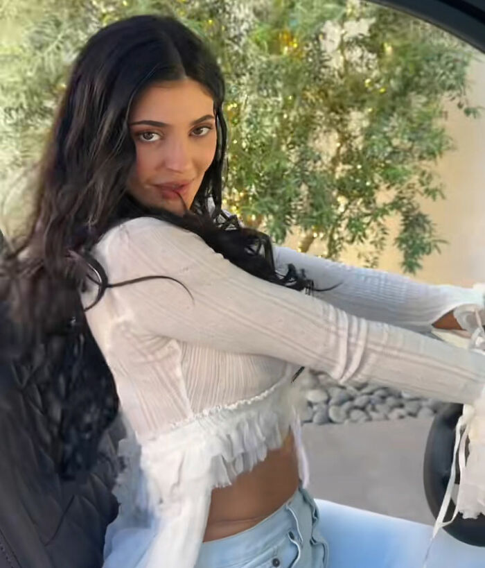 Kylie Jenner sentada com uma blusa branca e jeans com longos cabelos escuros durante a turnê da casa Coachella, provocando reação em massa.