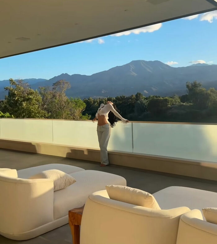 Mulher em pé na varanda com vista para as montanhas durante o passeio pela casa de Kylie Jenner Coachella com móveis modernos de exterior visíveis.