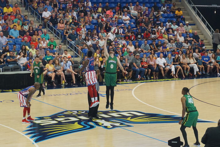 Basketball players on a court, one shooting, others watching. Spectators in the stands. God Bless America, once rich.