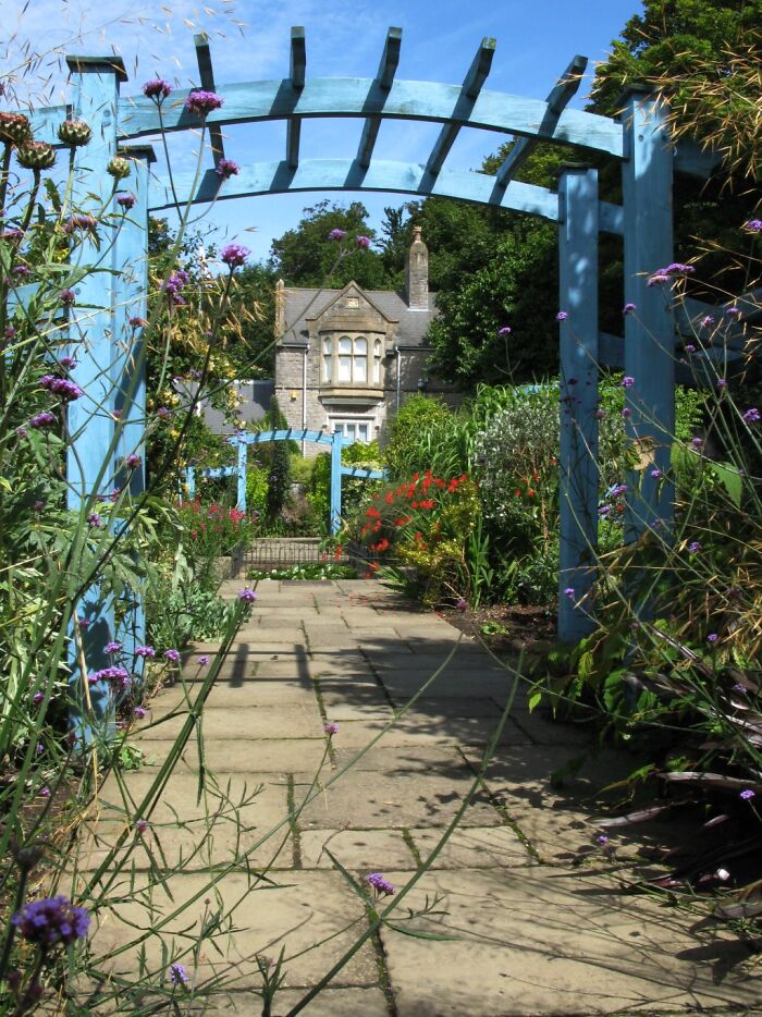 Blue garden archway over stone path with colorful flowers leading to an old stone house, evoking mysterious and creepy feelings.