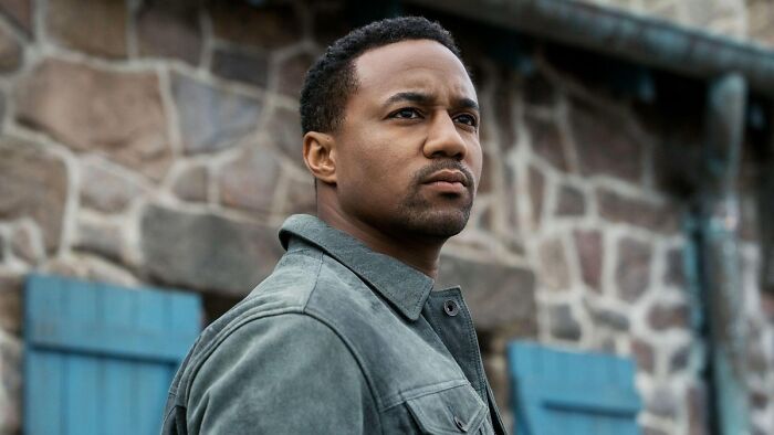 Actor portraying A-Train in The Boys Season 5, standing outside with a serious expression near blue window shutters.