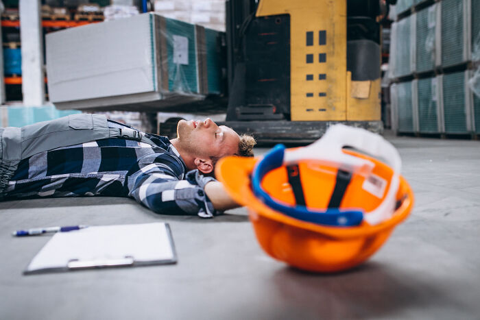 A worker lies unconscious on the floor next to an orange hard hat, highlighting workplace incidents and safety concerns.