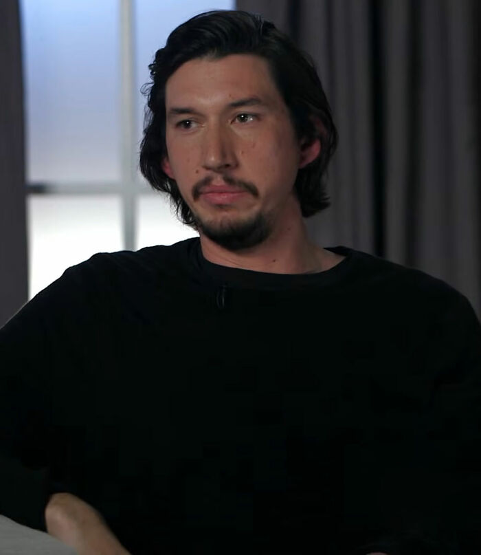 Adam Driver sitting indoors in a black shirt, looking pensive during an interview about Girls set accusations.