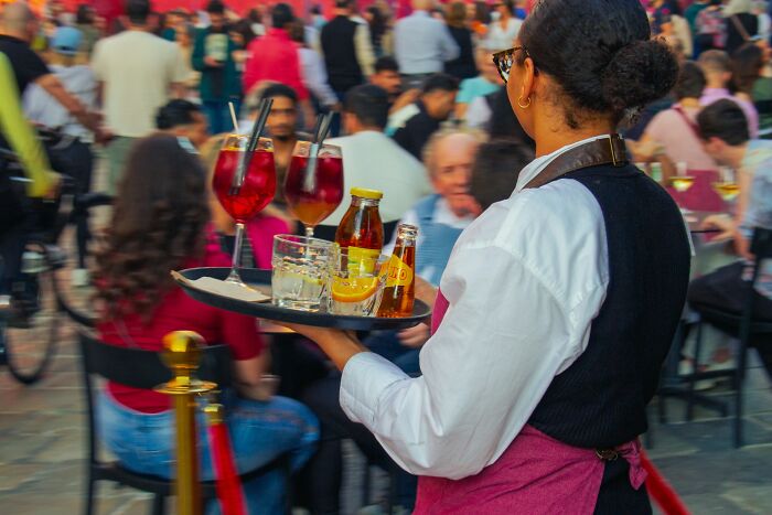 A waitress in a busy outdoor cafe carries a tray with drinks, a scene that might make some rage quit their jobs.