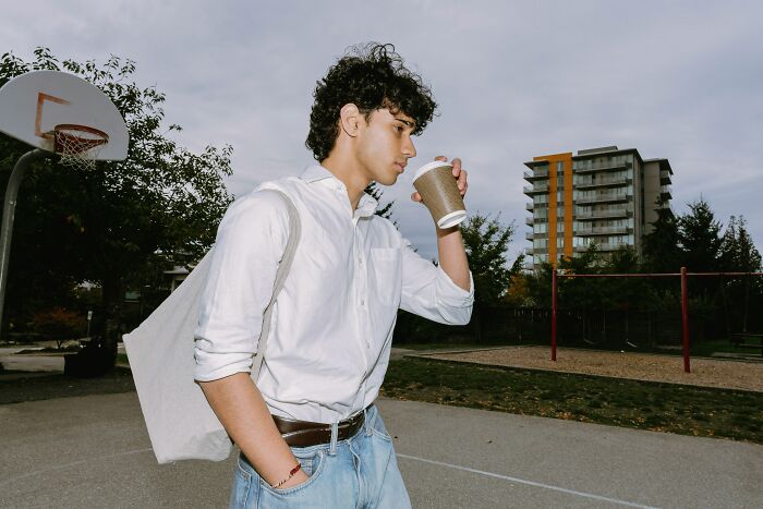 Young man displaying intelligence by carrying a coffee cup and tote bag while walking near a basketball court outdoors.