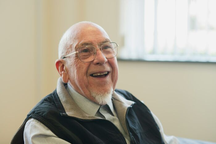Smiling elderly man wearing glasses and a black vest, enjoying a moment in a bright indoor setting.
