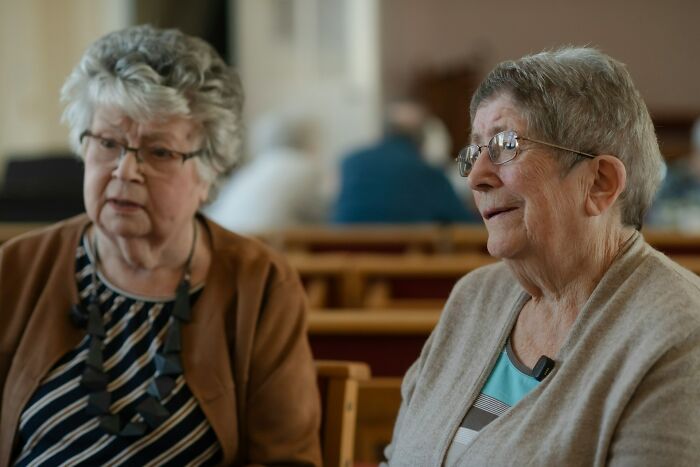 Two elderly women engaged in conversation at a wedding, witnessing signs of a future breakup among guests.
