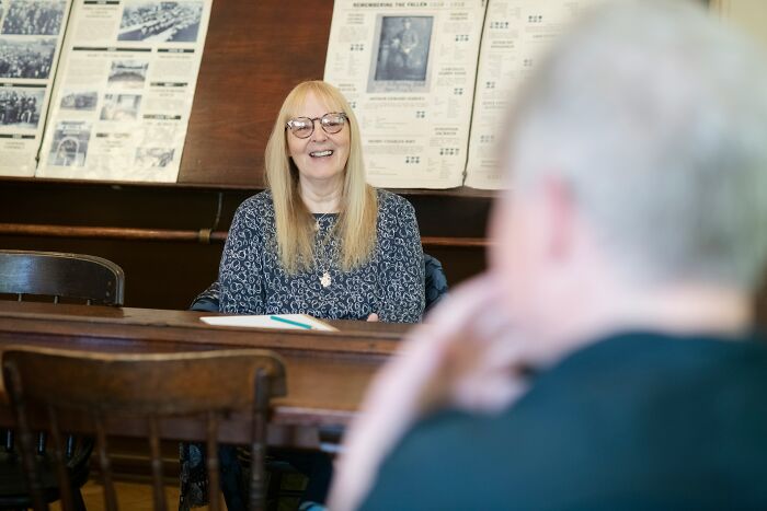 Woman with glasses smiling in a classroom setting, showing intelligence in a casual and engaging conversation.