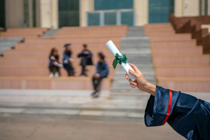 Graduate holding diploma in hand, with blurred graduates sitting on outdoor steps, highlighting random life decisions.