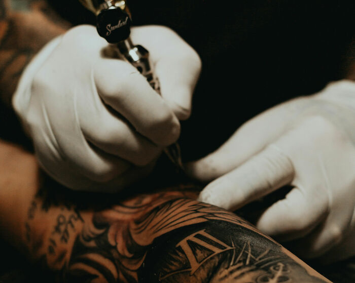 Close-up of a tattoo artist using a machine on an arm covered with multiple tattoos in a dimly lit setting. Close-up of a tattoo artist using a machine on an arm covered with multiple tattoos in a dimly lit setting.