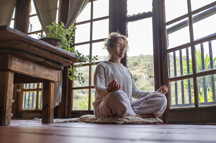 A person with curly hair meditating in a relaxed pose by large wooden windows, embodying cheat codes for surviving adult life.