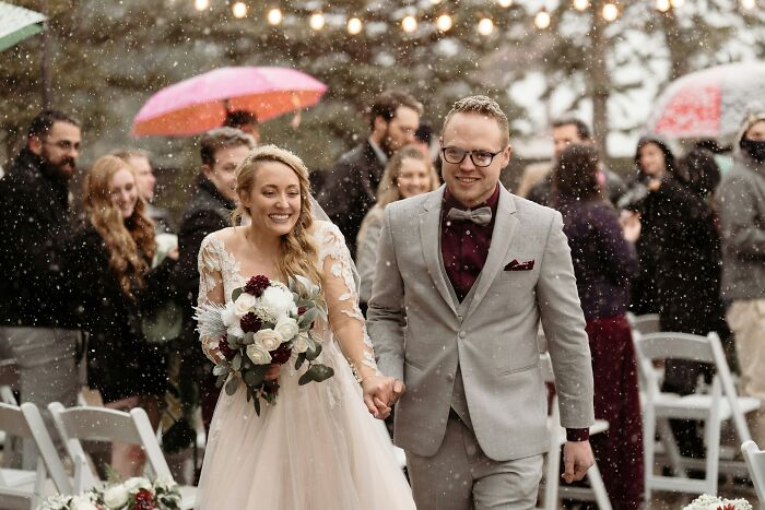 Bride and groom smiling and walking hand in hand during a snowy outdoor wedding with guests under umbrellas in the background.