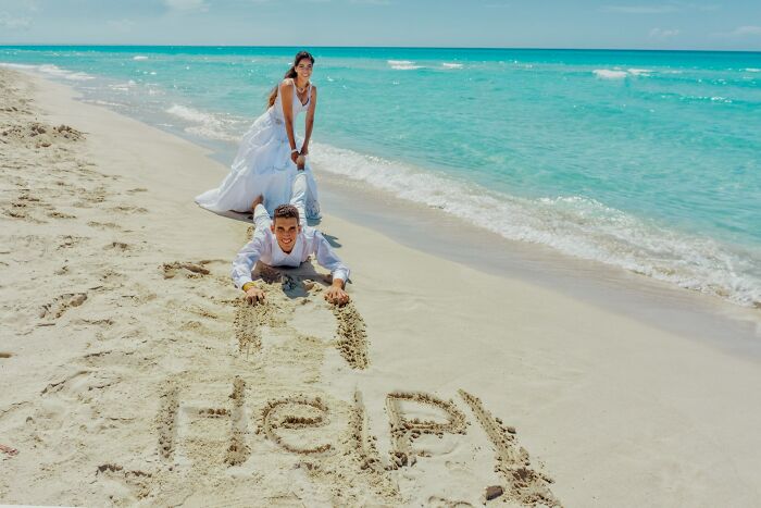 Bride in white dress dragging groom on sandy beach with the word help written in sand, illustrating trashy wedding stories.
