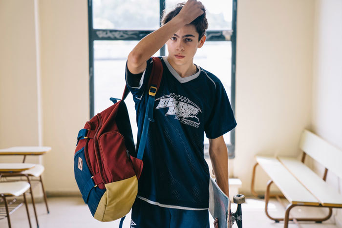 Young man in a sports jersey with a backpack, looking thoughtful in a classroom, highlighting challenges with autistic brother care. Young man in a sports jersey with a backpack, looking thoughtful in a classroom, highlighting challenges with autistic brother care.
