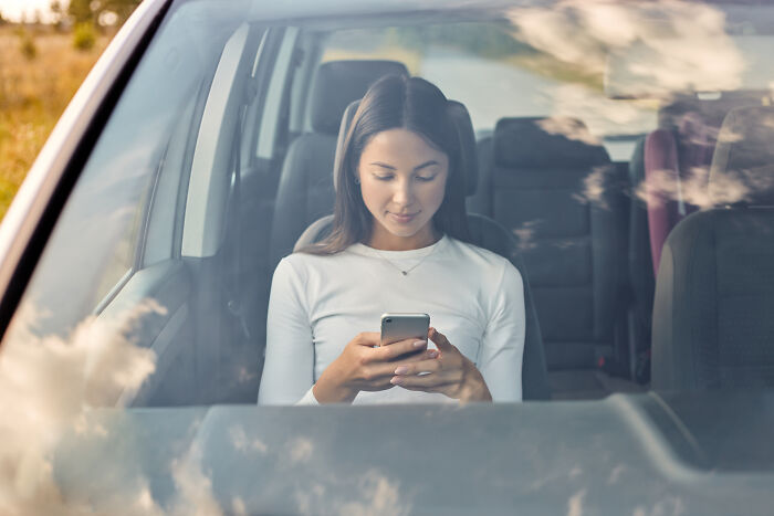 Young woman in a white shirt, sitting in a car and looking at her phone. She is having a horrible first date.