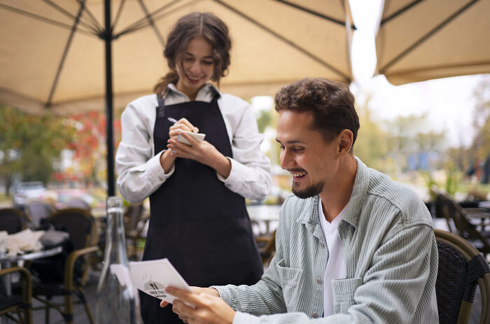 A man smiling at a woman, a waitress, who is taking his order at an outdoor cafe. Possibly a first date.