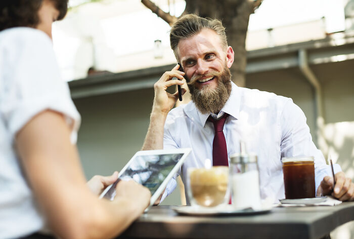 Smiling man with a beard on his phone at a cafe, possibly on one of his first dates. A person uses a tablet in the foreground.