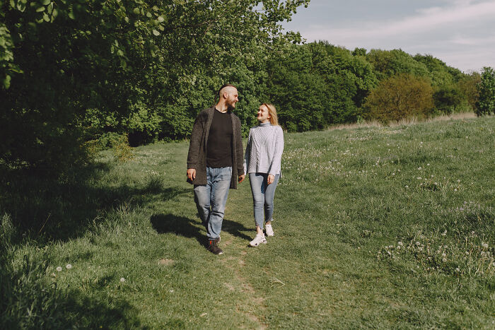 A smiling couple, dressed casually, walks hand-in-hand through a grassy field with trees in the background. Their joyful expressions contrast with the idea of horrible first dates and character-building trauma.