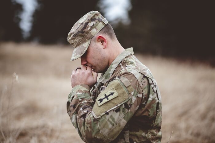 A man in military camouflage uniform, head bowed, hands clasped, appearing to pray or reflect. Could this be one of those horrible first dates?