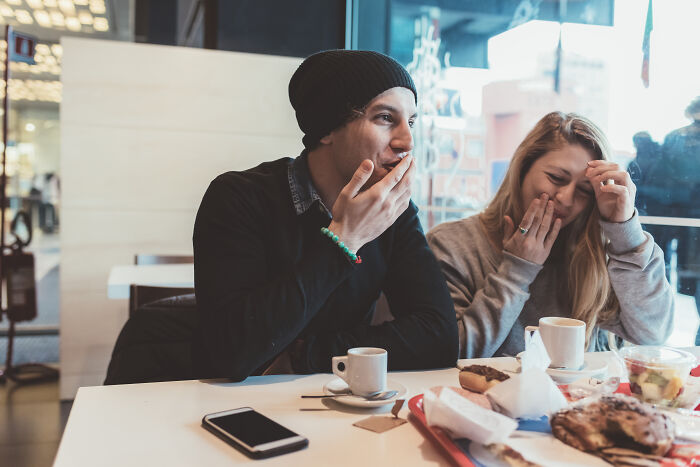 A man in a black beanie and a woman laugh, covering their mouths, on a first date. This might be a horrible first date.