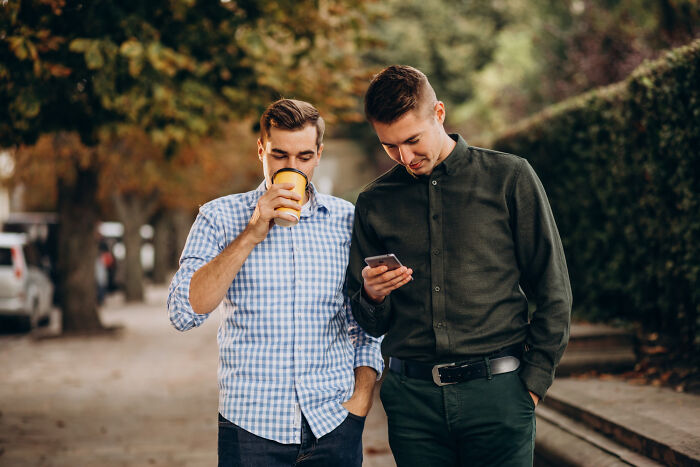 Two men walking outdoors, one drinking coffee and the other checking his phone, illustrating real life plot twists.