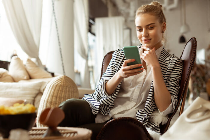Young woman sitting comfortably at home, smiling while reading surprising real life plot twists on her phone.