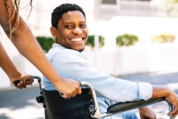 Smiling man in wheelchair outdoors with woman pushing, illustrating real life plot twists involving family relationships.