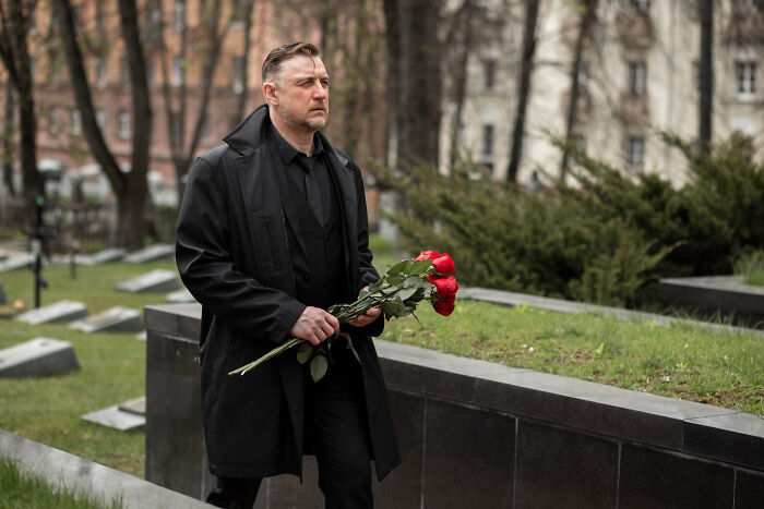 Man in black coat holding red roses at a cemetery, representing real life plot twists involving family relationships.