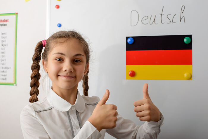 Young girl in a classroom giving thumbs up with a German flag and the word Deutsch on the whiteboard behind her.
