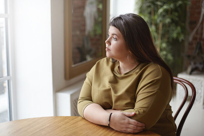 A woman in a green shirt sitting at a wooden table, looking thoughtfully out a window, reflecting on real life plot twists.