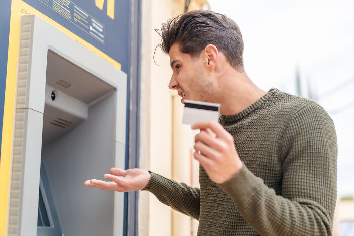 Young man at ATM holding card with confused expression, illustrating real life plot twists involving family relationships.