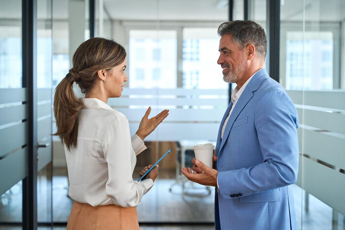 Two coworkers in business attire discussing surprising real life plot twists during a casual office meeting.