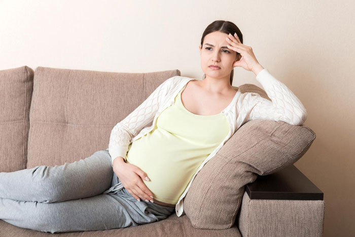 Pregnant woman sitting on couch looking frustrated while holding her belly, reflecting stress about raising her baby.