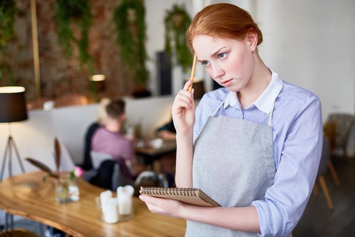 Young book store manager in apron holding a notebook and pencil, appearing thoughtful in a cozy café setting. Young book store manager in apron holding a notebook and pencil, appearing thoughtful in a cozy café setting.