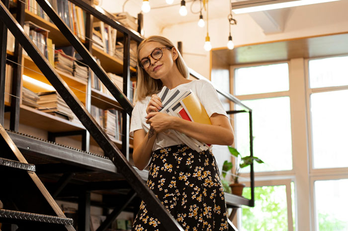 Young book store manager holding books near shelves, smiling confidently in bright modern bookstore setting. Young book store manager holding books near shelves, smiling confidently in bright modern bookstore setting.