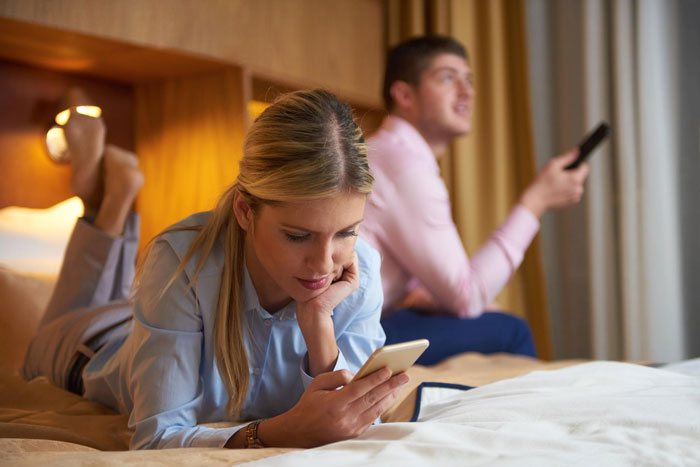 Young woman looking confused at her phone while her boyfriend watches TV, highlighting connection and relationship problems.