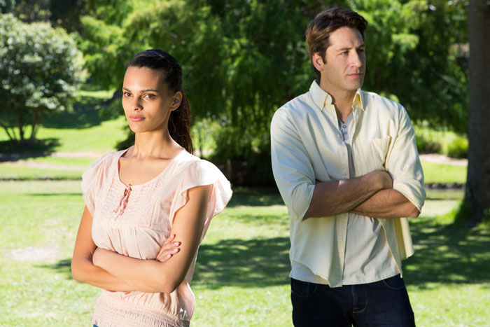 Couple standing apart with arms crossed in a park, showing tension and conflict during a birthday party scene.