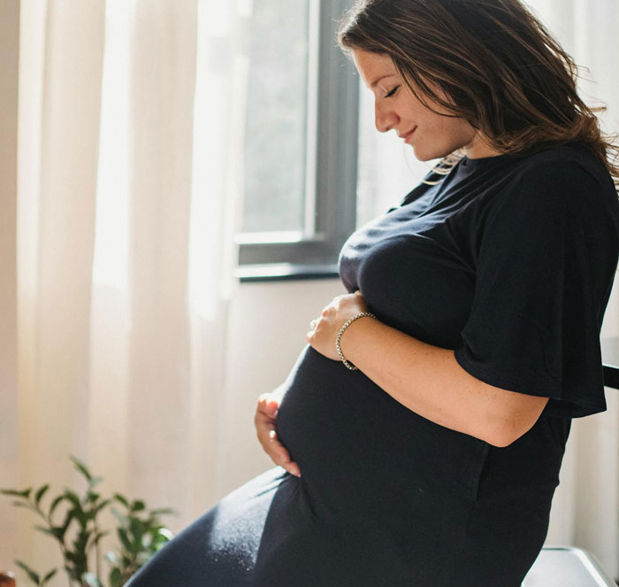 A woman with a baby bump, dressed in black, holds her belly and smiles softly, bathed in natural light.